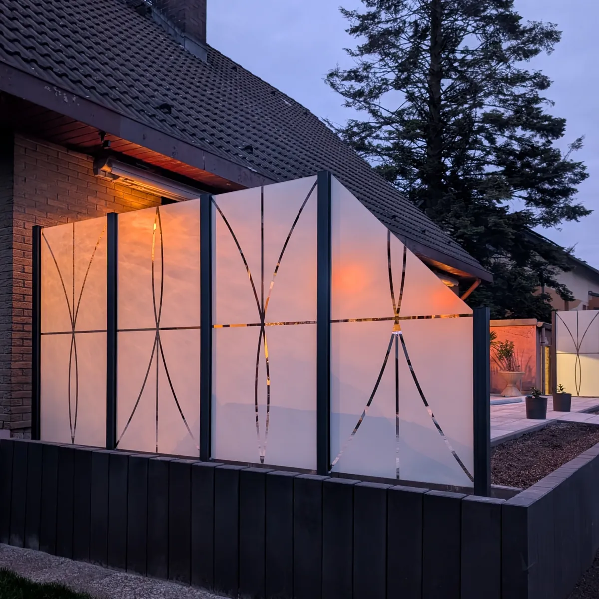 Glaszaun mit dem Motiv Elipsa am Abend, hinter dem Glas brennt warmes Licht